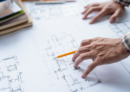 A person sketches on a blueprint with a pencil, focusing intently on the detailed design.