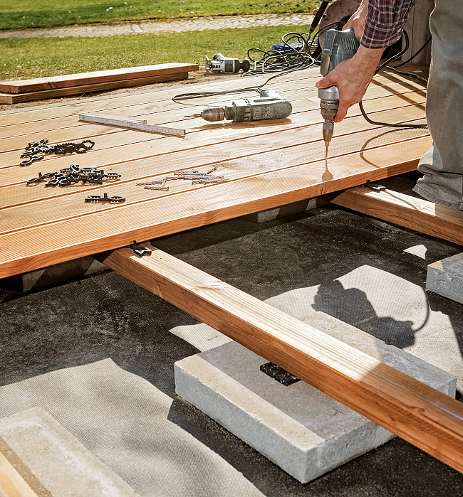 A man operates a drill while constructing a wooden deck in a residential outdoor setting.